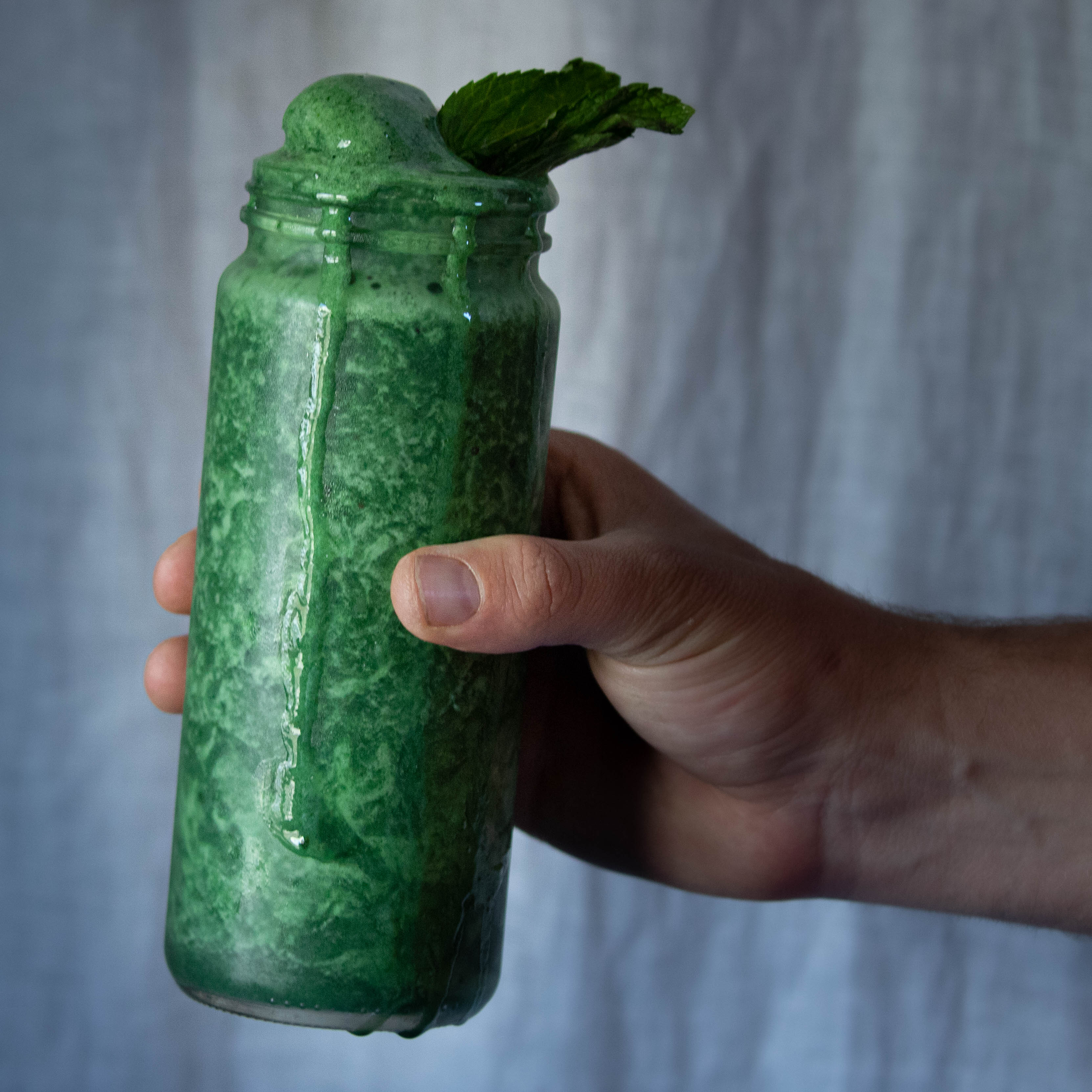 A frosty glass overflowing with a green, minty smoothie is held in hand in front of a white linen backdrop