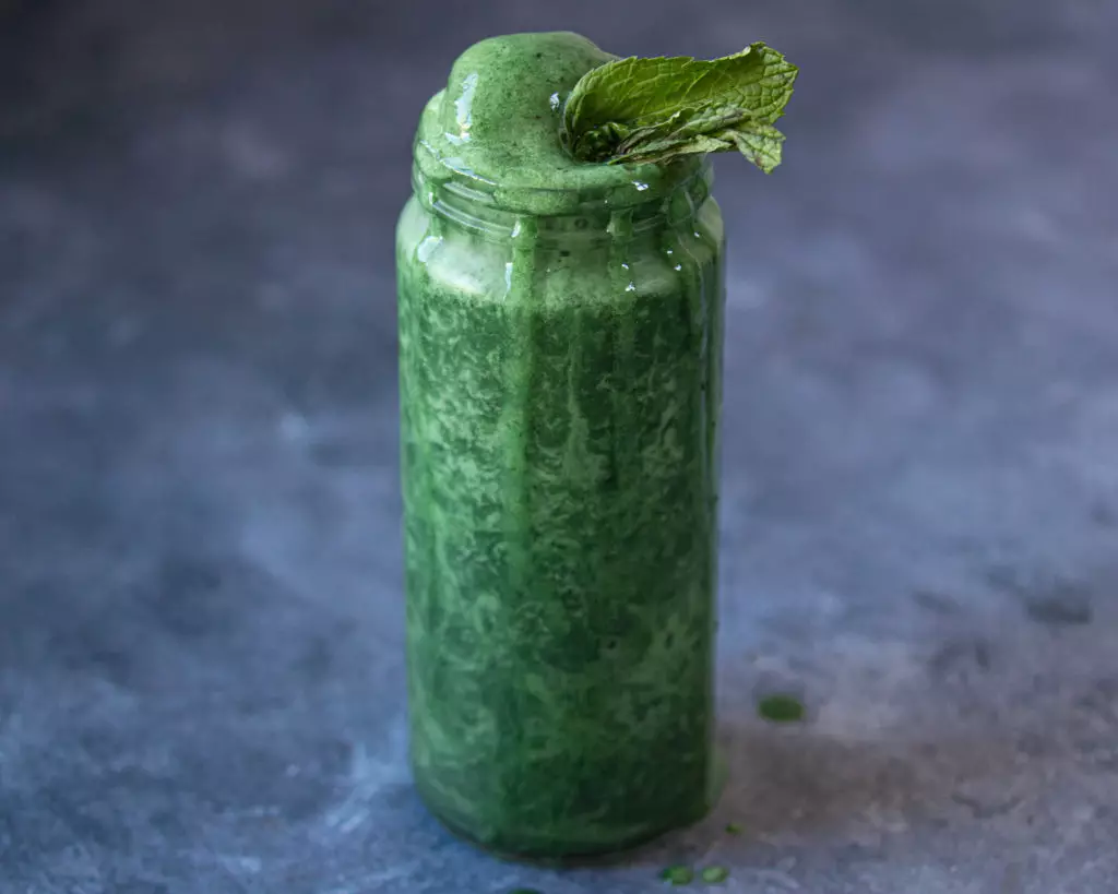 Tall glass of a green smoothie with drips down the side of the glass and mint leaves on top