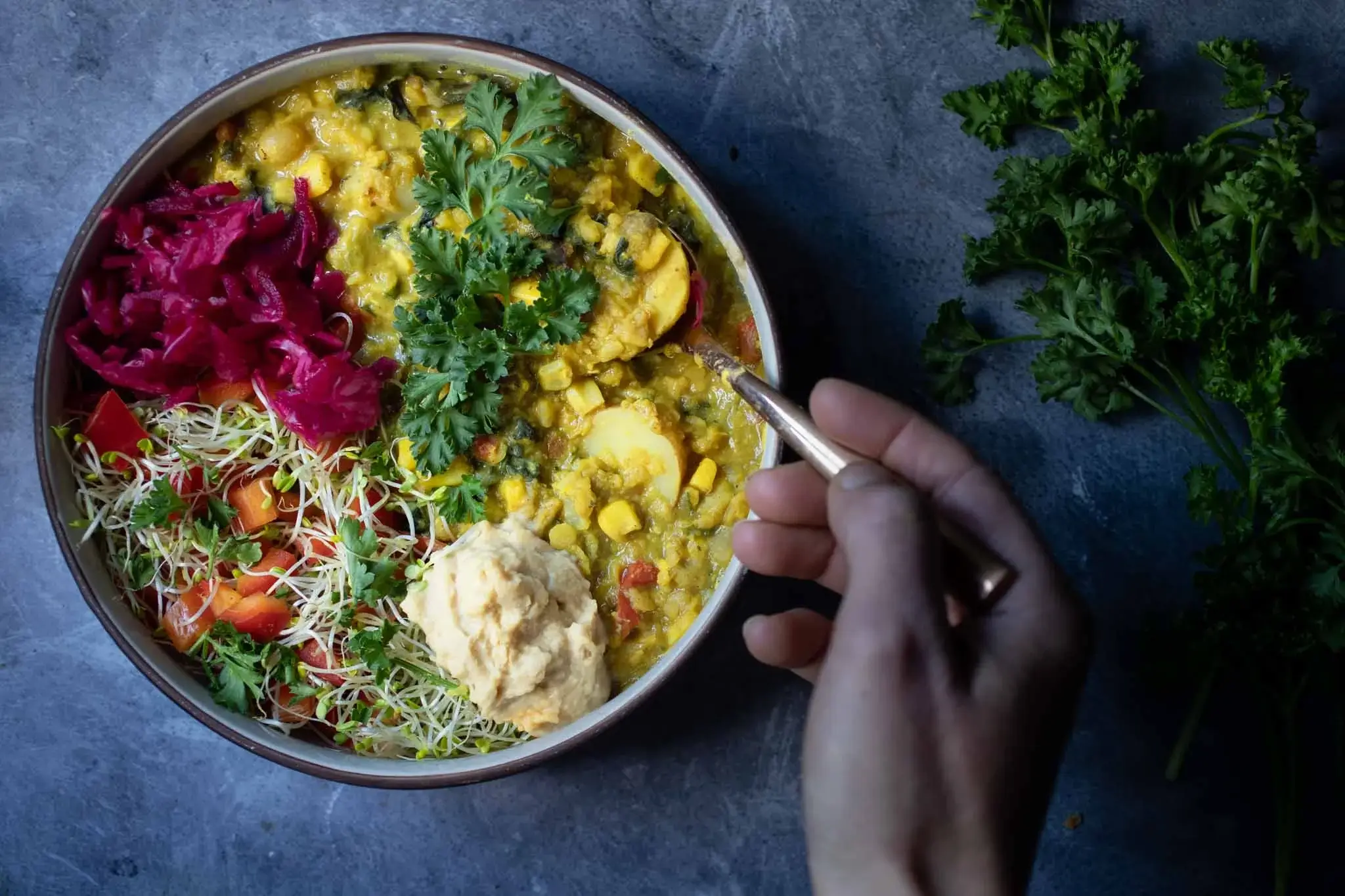 a hand stirs a bowl of lentil stew garnished with parsley and served with hummus, sauerkraut, and sprouts